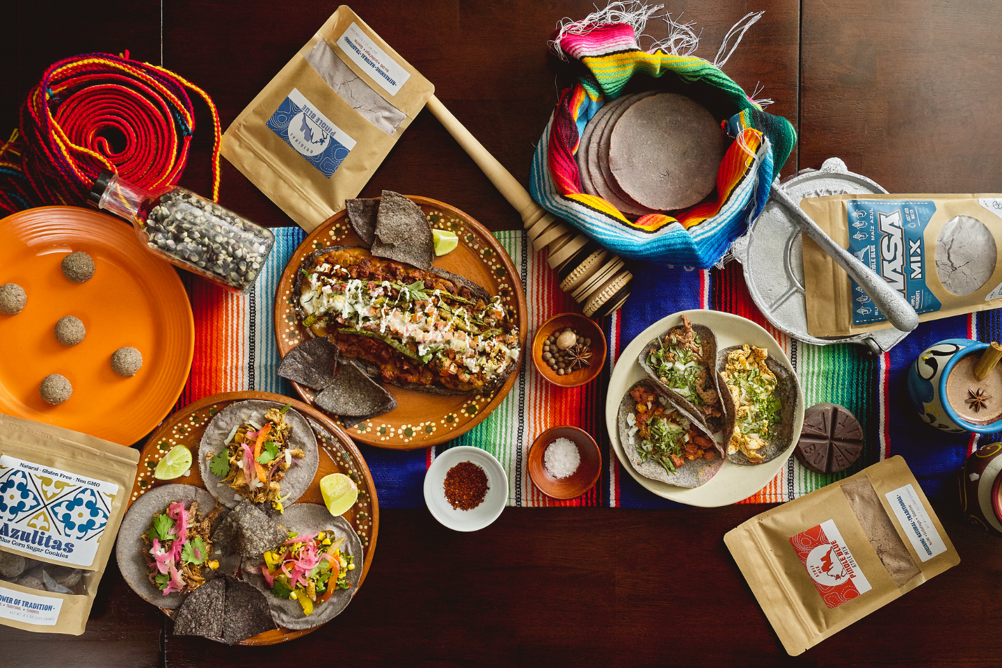 Table with lots of hispanic food on it, including tortillas, tacos, and cookies. Pinole Blue product packages are displayed as well.